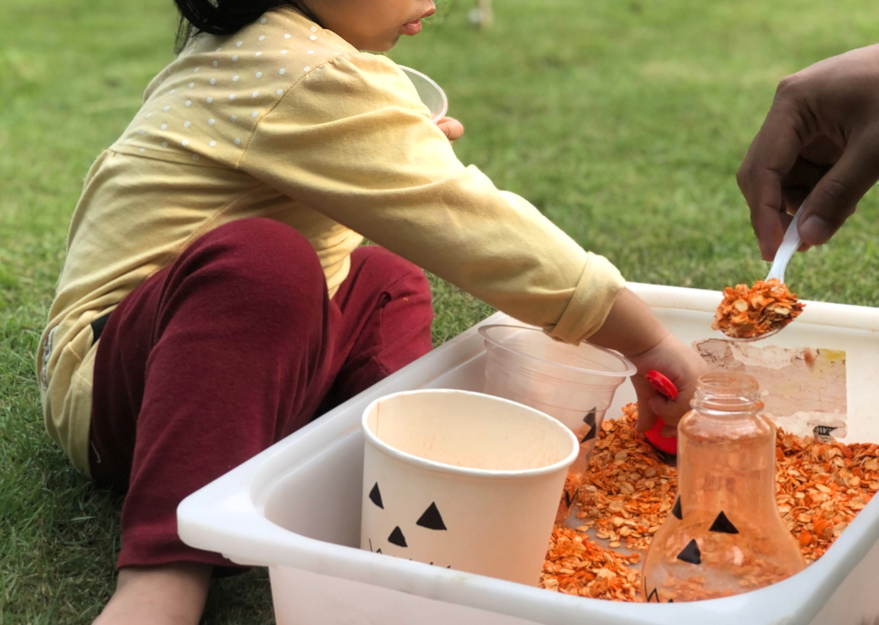 A child and an adult scooping dried corn together in an outdoor sensory bin