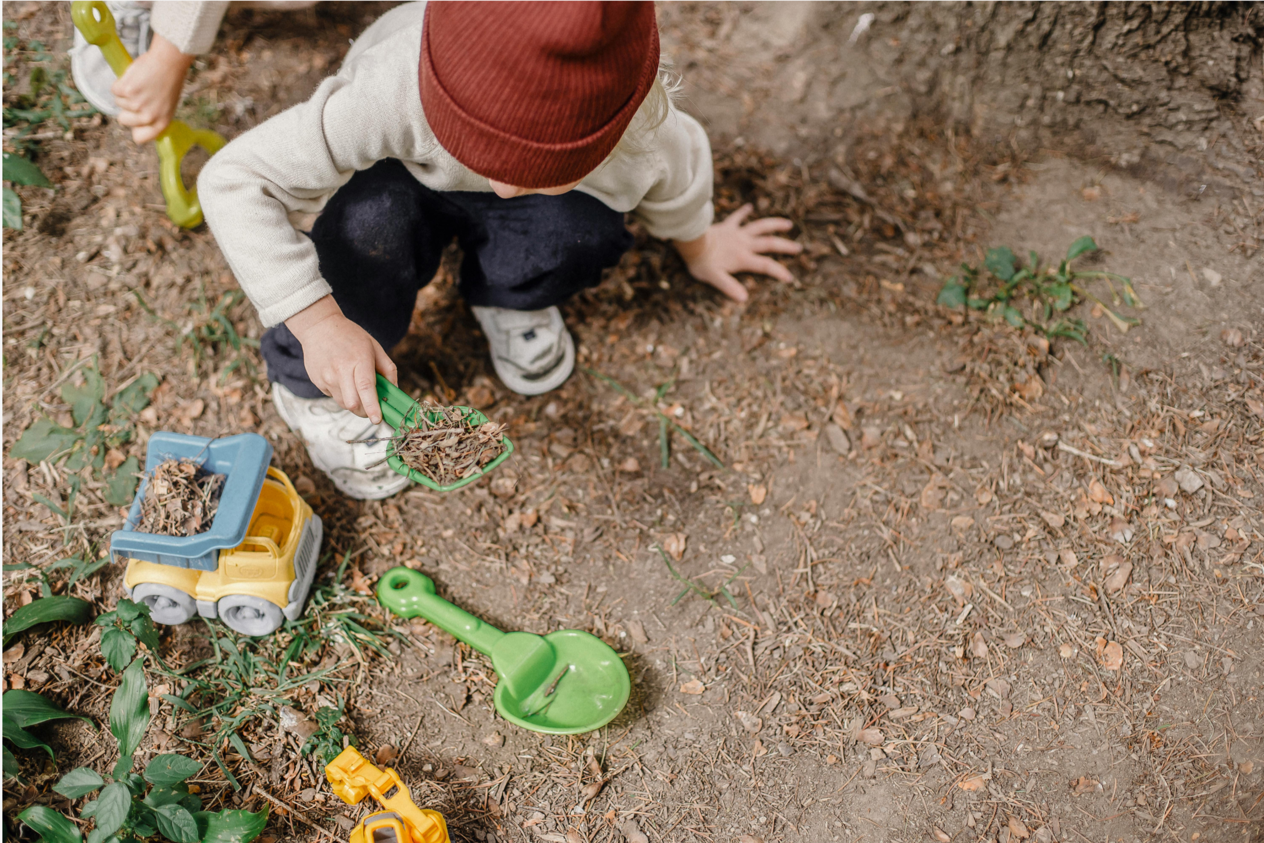 A child in a red beanie crouching on the ground, digging with a toy spade and truck